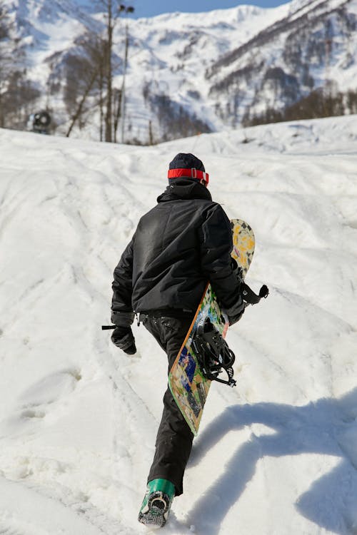 A Person in Black Jacket and Pants Walking on a Snow Covered Ground while Carrying a Snowboard