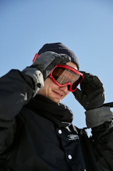 Adult man smiling while adjusting goggles in winter attire against a clear blue sky.