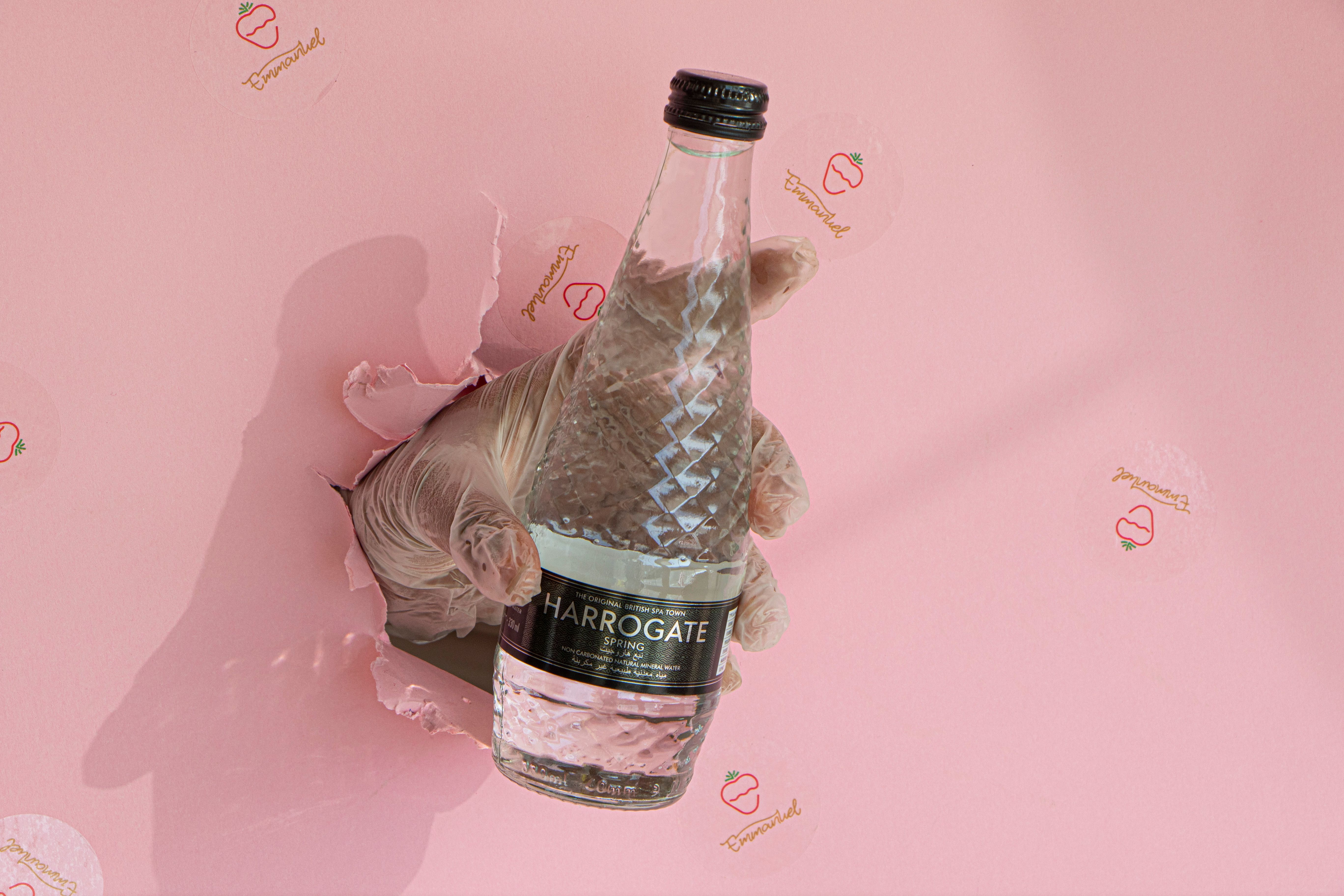 Gloved hand holding Harrogate water bottle through torn pink paper background.