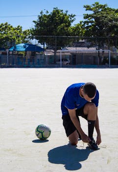 A soccer player in blue shirt ties his shoelace on an outdoor field.