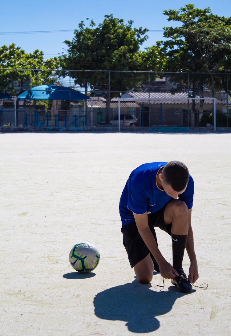 Man In Blue Shirt Tying His Shoelace On Soccer Field