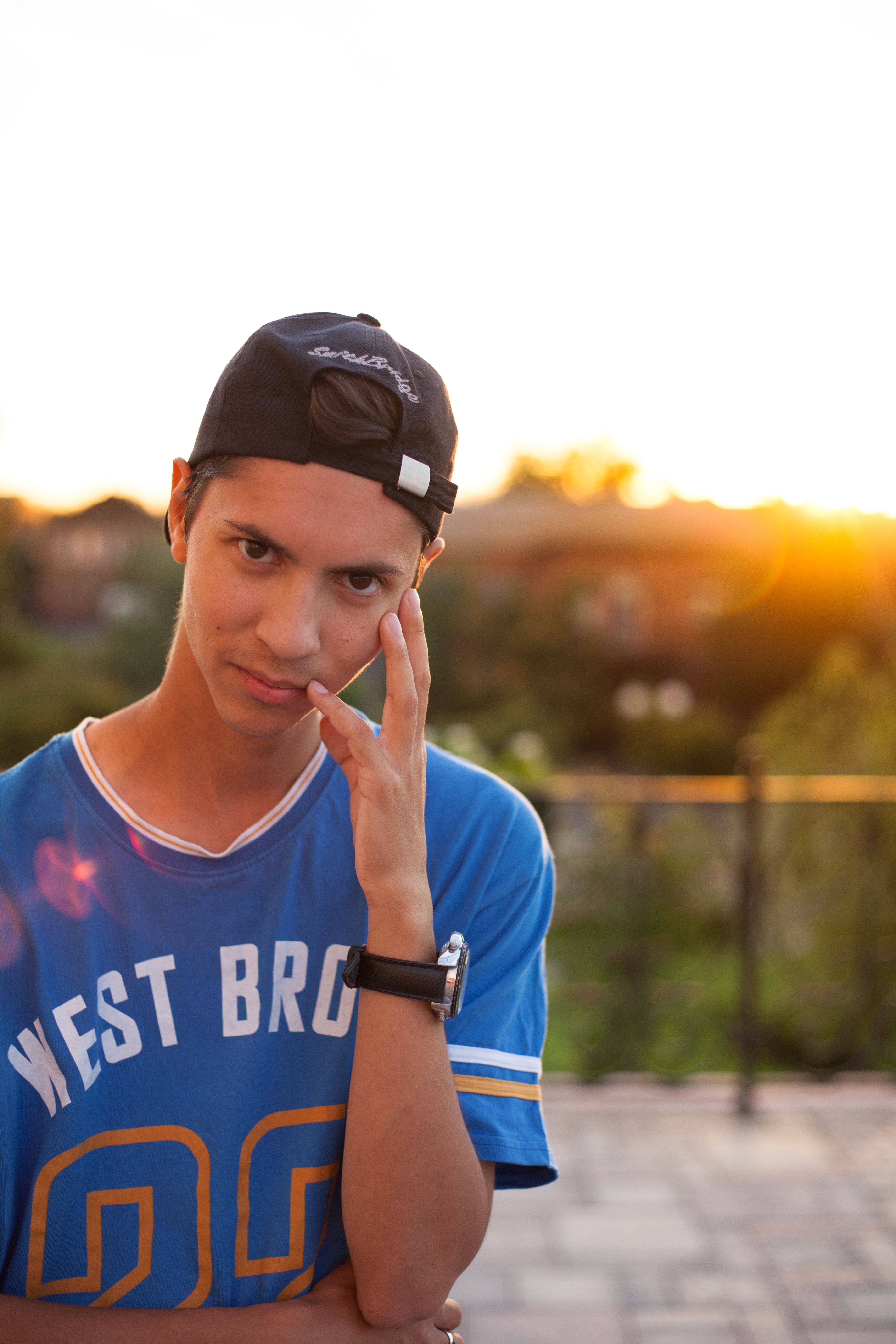 A teen wearing a black cap and blue shirt outdoors with a blurred sunset background.
