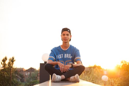 Free A young man wearing a blue shirt and cap sits on a ledge outdoors at sunset, exuding a relaxed vibe. Stock Photo