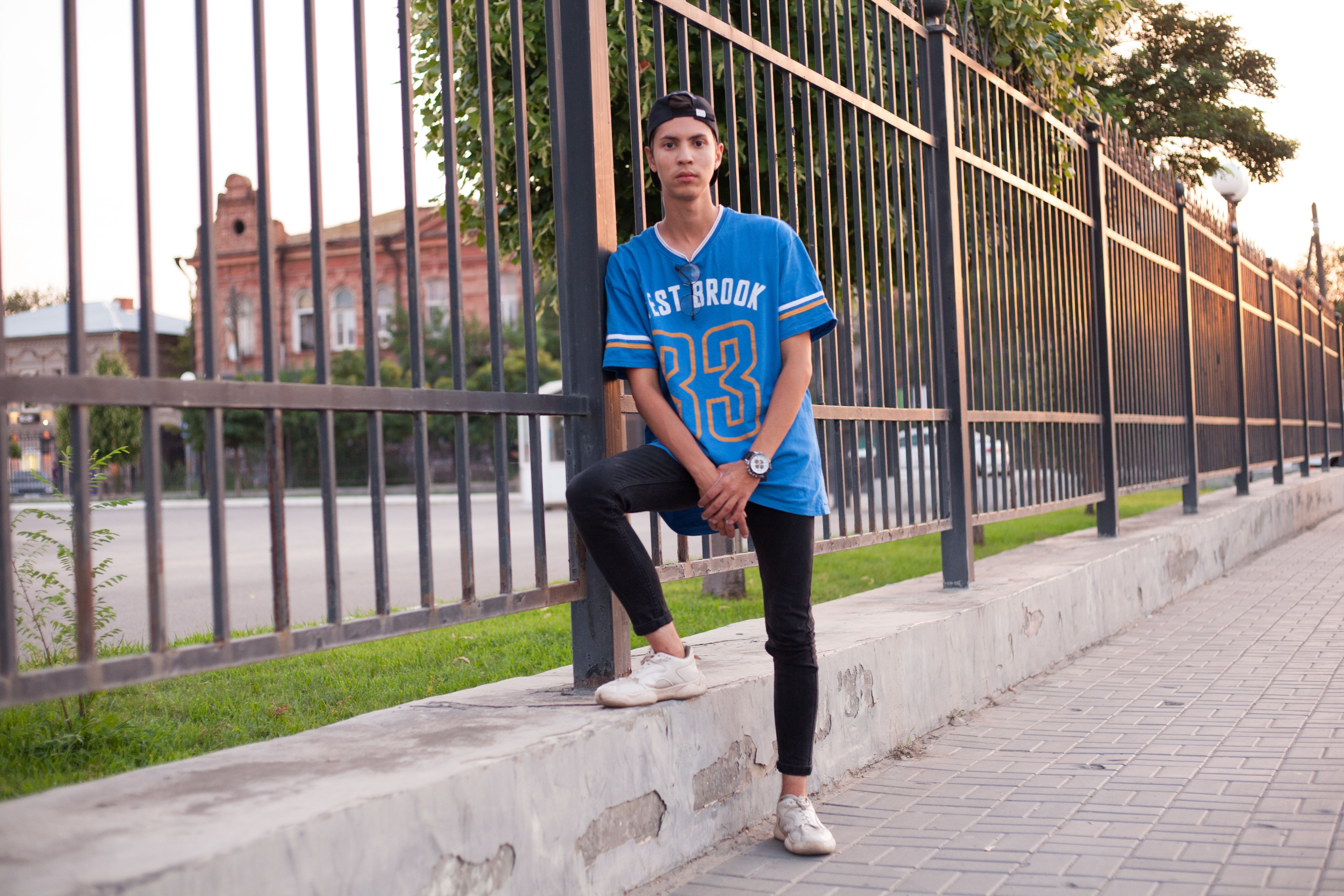 Young man standing by a metal gate in a blue sports jersey, outdoor urban scene.