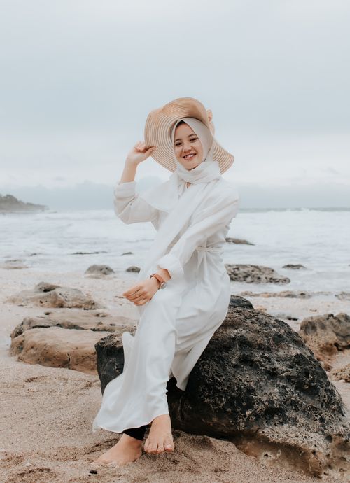 Free A woman in a white dress and hat smiles while sitting on a rock by the sea. Perfect for travel and lifestyle themes. Stock Photo