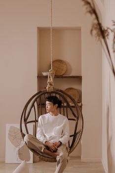 Asian man in a white sweater sits relaxed in a hanging chair indoors, exuding a calm and serene atmosphere.