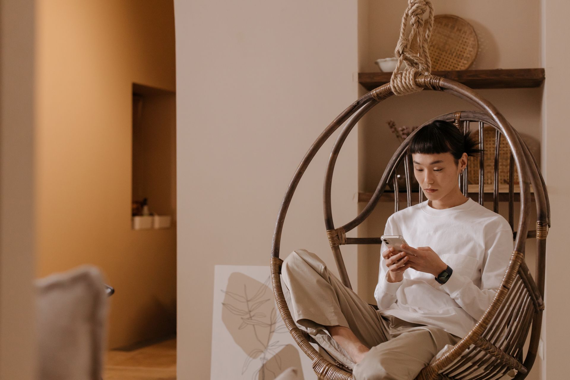 Asian man sitting in a hanging chair, texting on a smartphone indoors, wearing a white sweater.