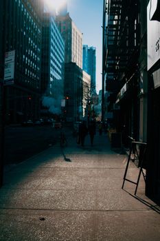 A dynamic city street with towering buildings and pedestrians in silhouette under the sun.