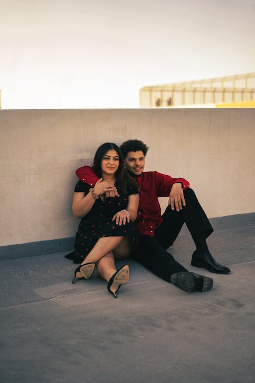 Full body of positive romantic Indian couple in stylish outfits hugging while sitting near concrete barrier of building against cloudless sky
