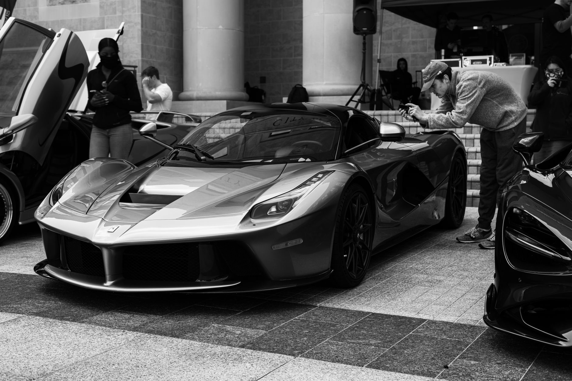 Black and white image capturing a luxury sports car at an outdoor car show with people interacting.