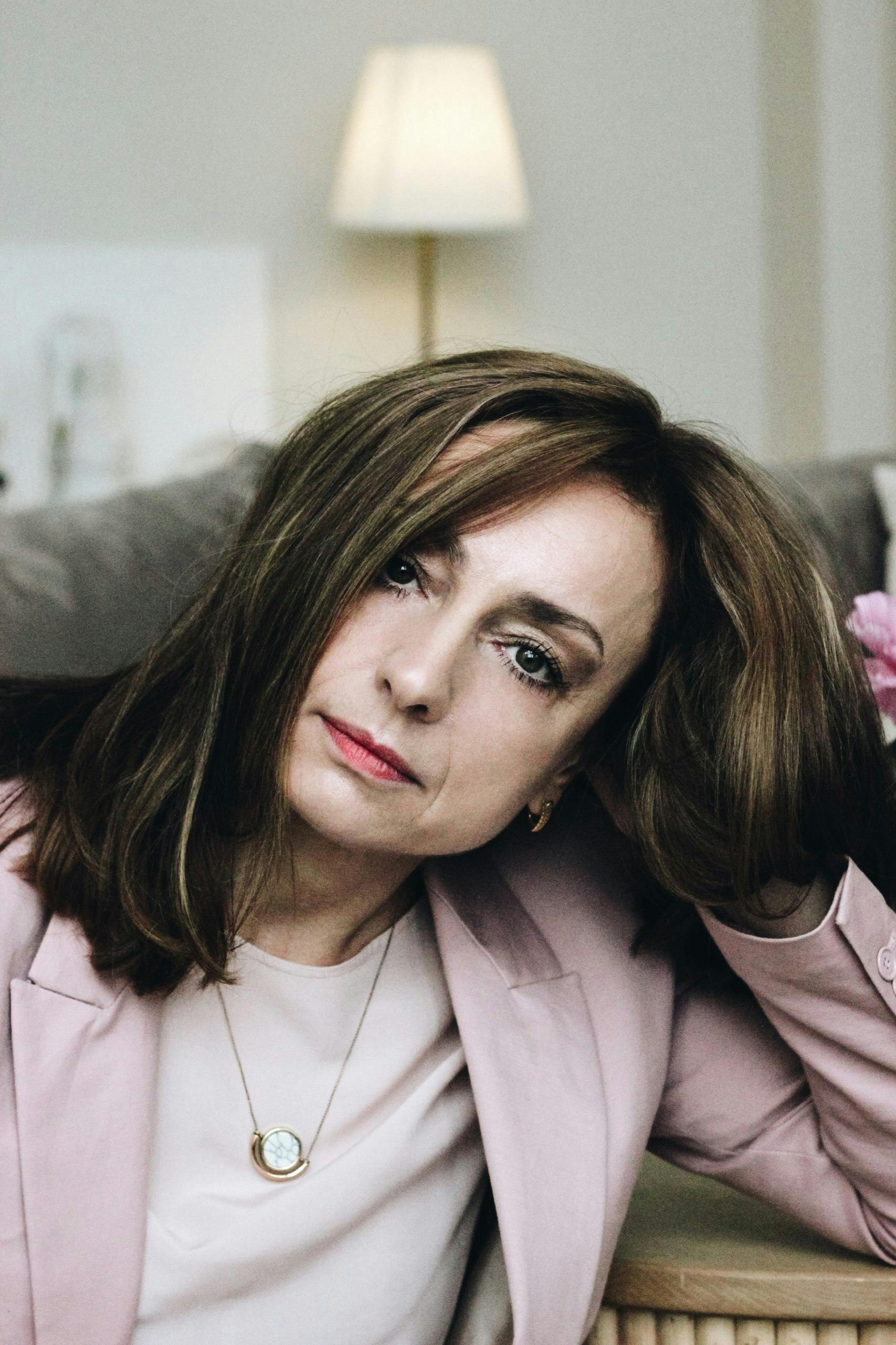 Stylish woman in a pink jacket with red lipstick indoors, gazing thoughtfully.