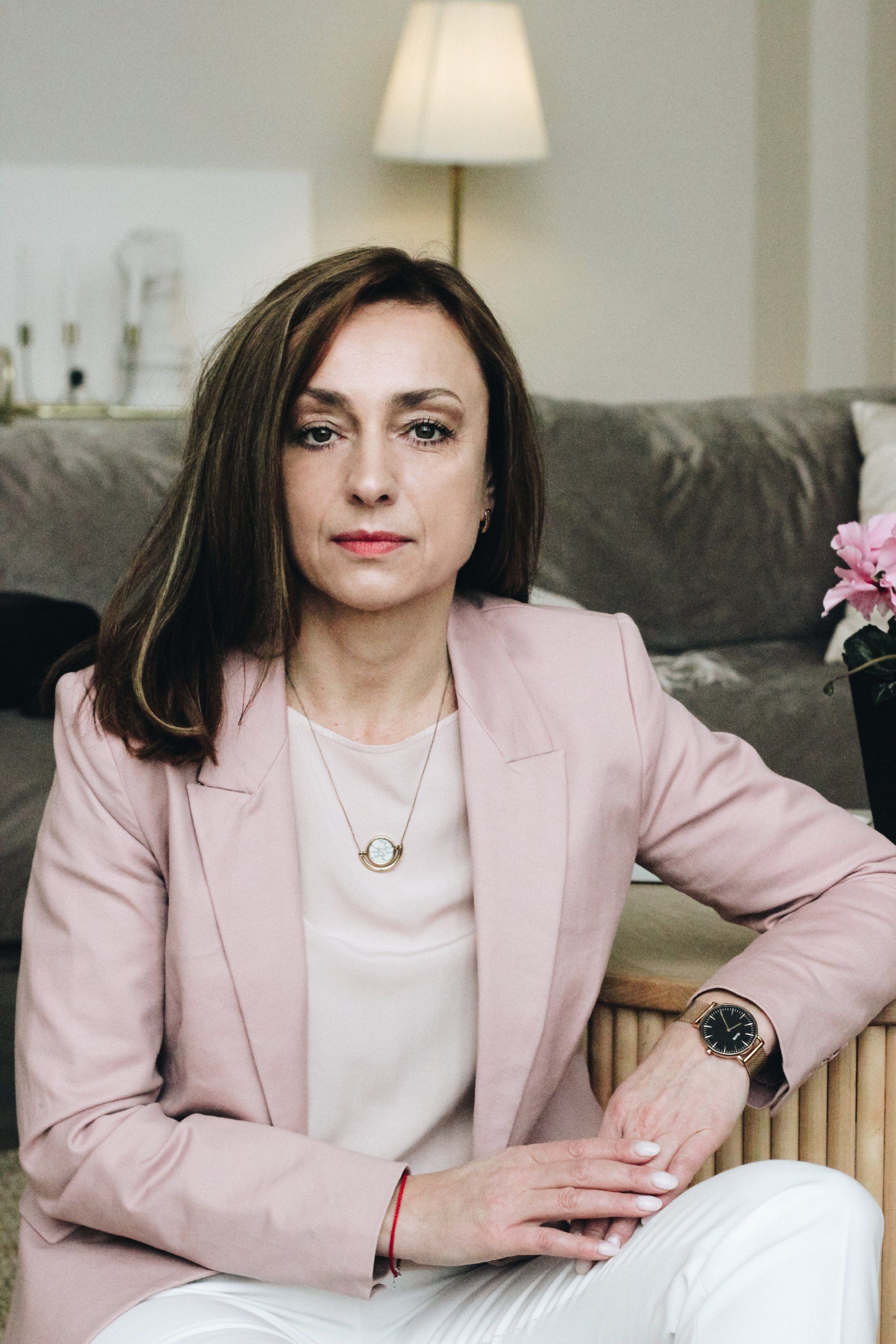 Elegant woman in business attire with pink suit and pendant, seated indoors.