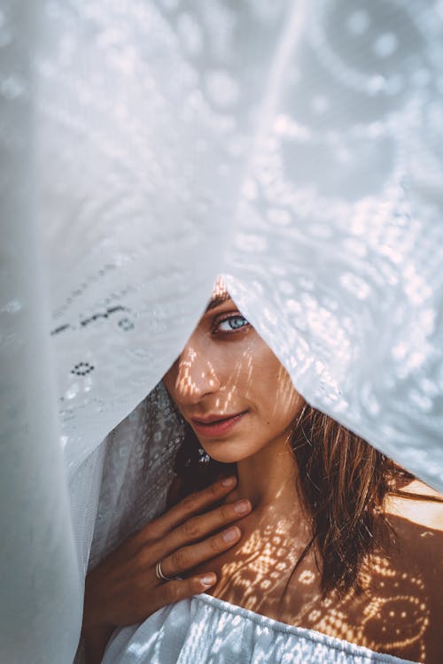 Headshot of young female model looking at camera while hiding under curtain and showing ring on hand in summer in daylight