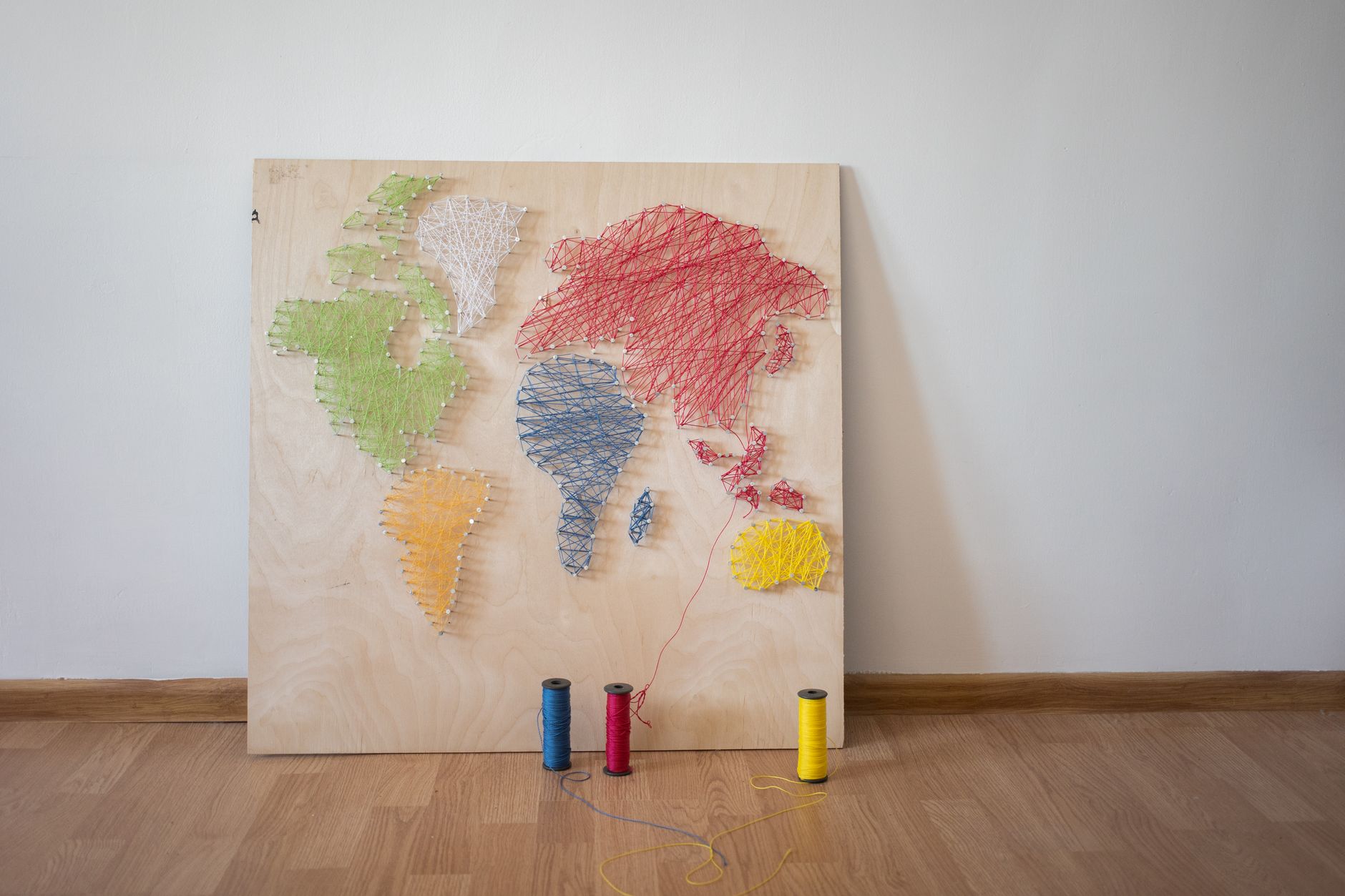 Board with string artwork of world map leaned on white wall near multicolored bobbins of thread on floor