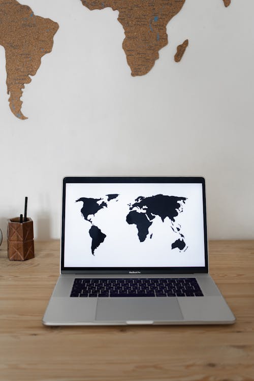 Laptop with black world map on screen placed on wooden table near white wall with silhouettes of continents