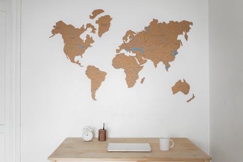 Alarm clock near pen container with cup and laptop on table placed near silhouettes of world map on wall