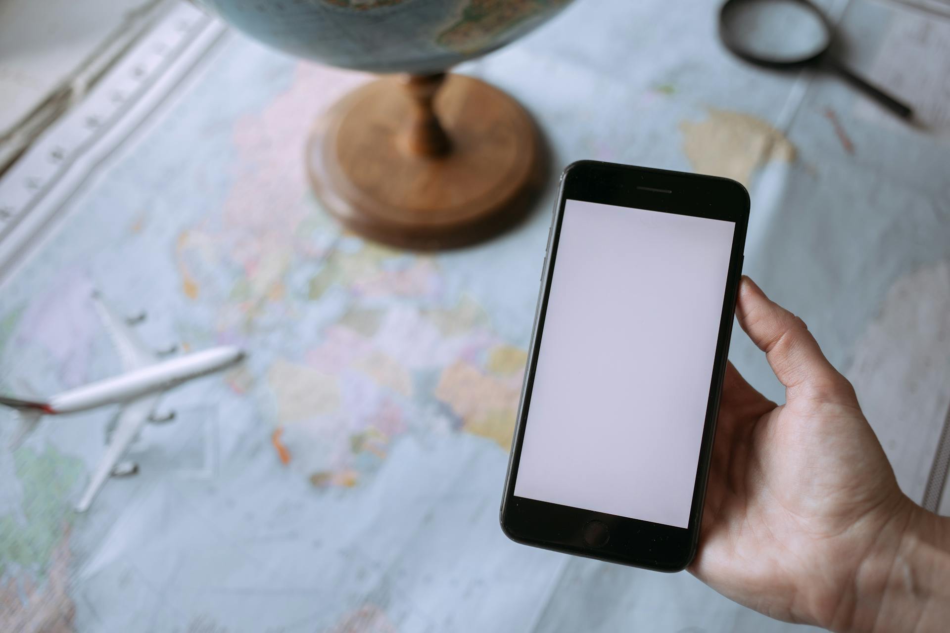 Hand holding smartphone over world map with globe and toy airplane, symbolizing travel planning.