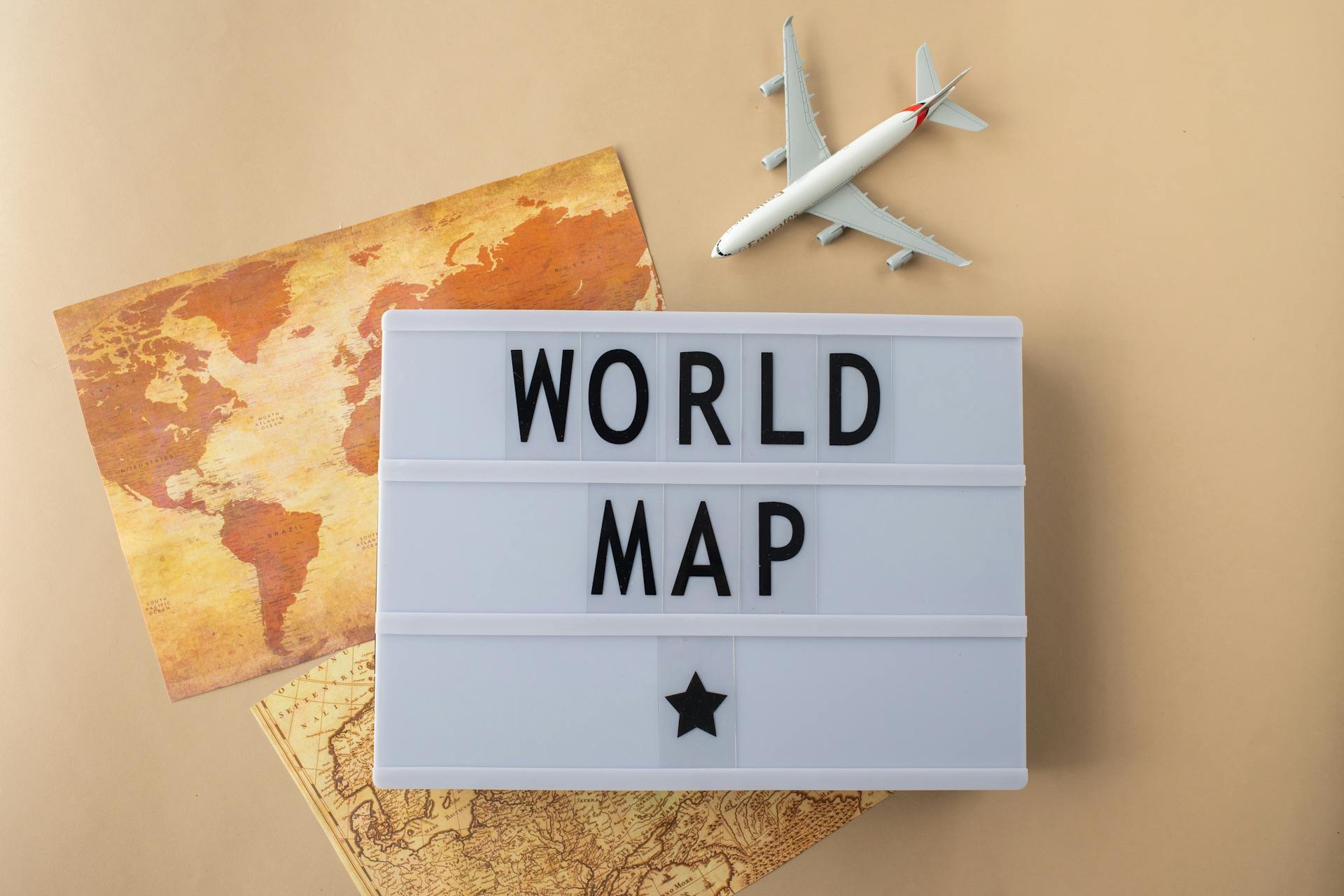 Top view of light box with World Map inscription placed near world continents and toy plane on beige background in studio