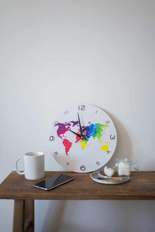 Round clock with colorful continents placed on wooden table with cup of beverage and cellphone near sugar on white background