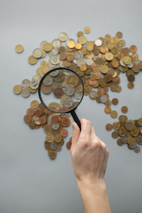 Top view of crop faceless traveler with loupe magnifying view of world map made of coins on gray background in light studio