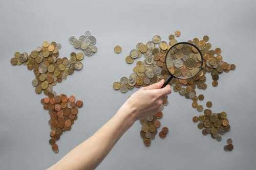 Free Top view of crop unrecognizable traveler with magnifying glass standing over world map made of various coins on gray background Stock Photo