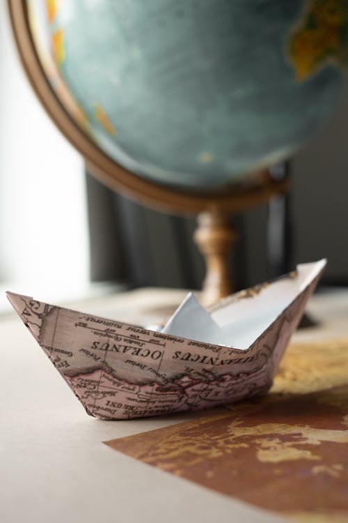 Paper boat made of world map placed on brown wooden table near round globe with continents in light room at home