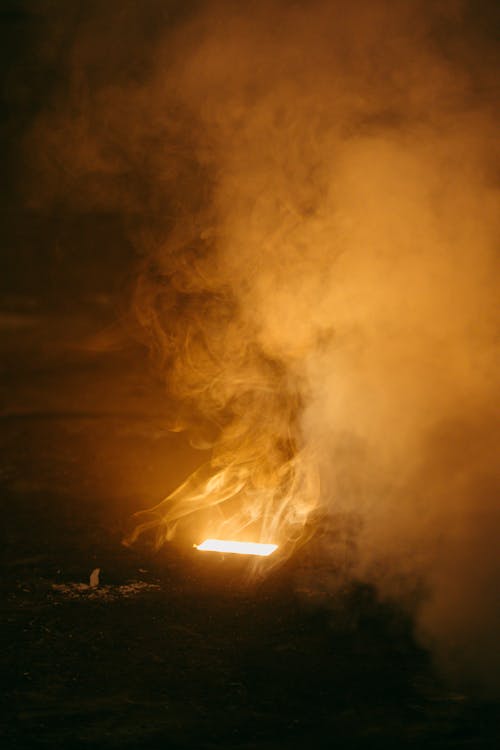 Side view of anonymous person sitting against bright flame under cloud diffusing in air at dusk