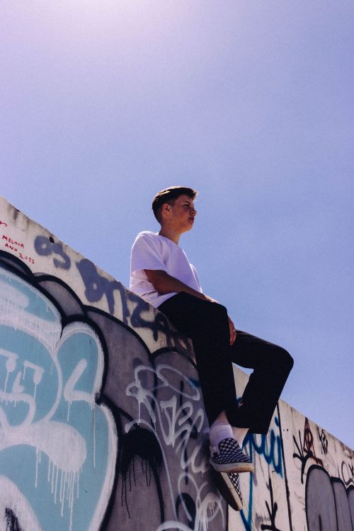 Free A young man sits atop a graffiti-covered wall under a clear sky, captured in Araranguá, Brazil. Stock Photo