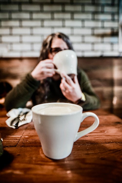 Free Anonymous female enjoying hot drink at wooden table with cup of coffee with milk foam in cafeteria Stock Photo