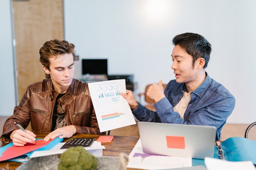 Two men discuss graphs and data at a shared workspace in a modern office setting.