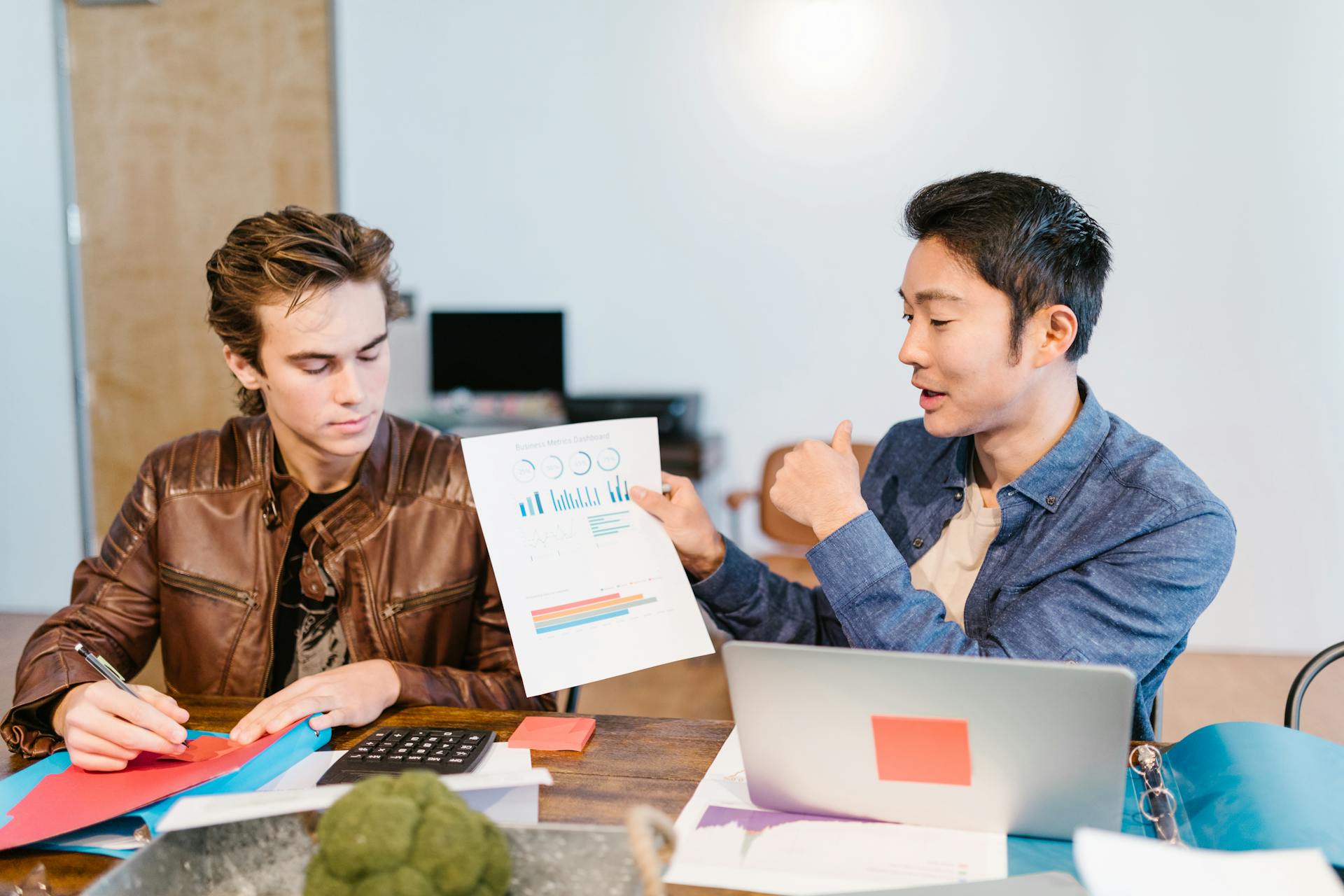 Two men discuss graphs and data at a shared workspace in a modern office setting.