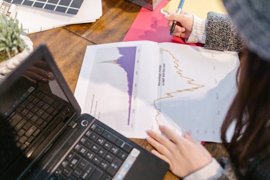 Person reviewing financial graphs and charts while taking notes beside a laptop.