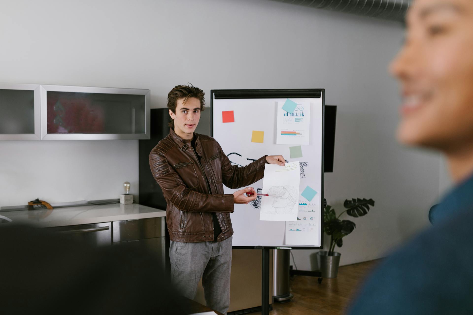 A man in a leather jacket presenting a business plan on a flip chart in a modern office setting.