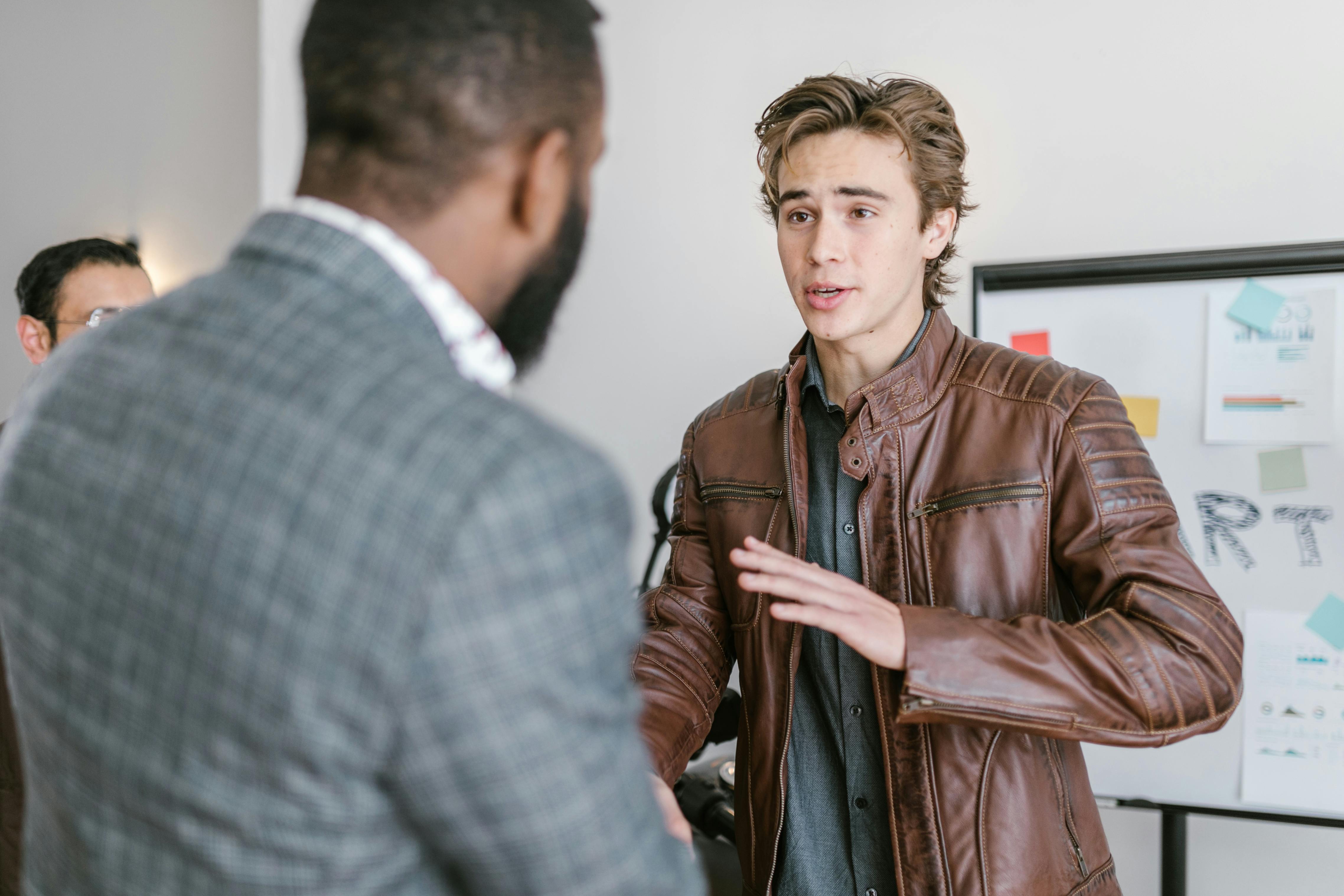 A young man in a leather jacket talking with colleagues during a business meeting indoors.