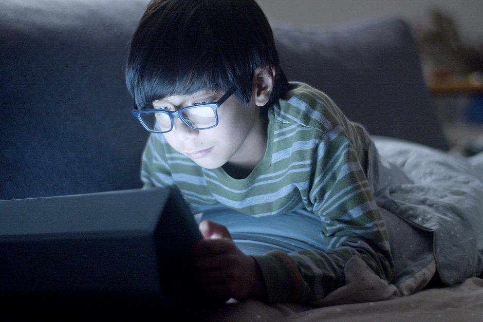 Young boy in a striped shirt using a tablet in bed at night with eyeglasses on.