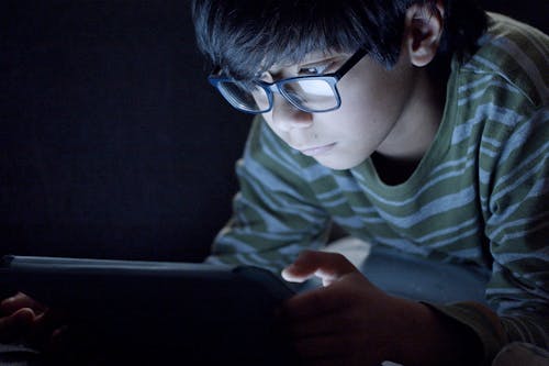 Free Boy Wearing Eyeglasses Using a Tablet in the Dark Room Stock Photo
