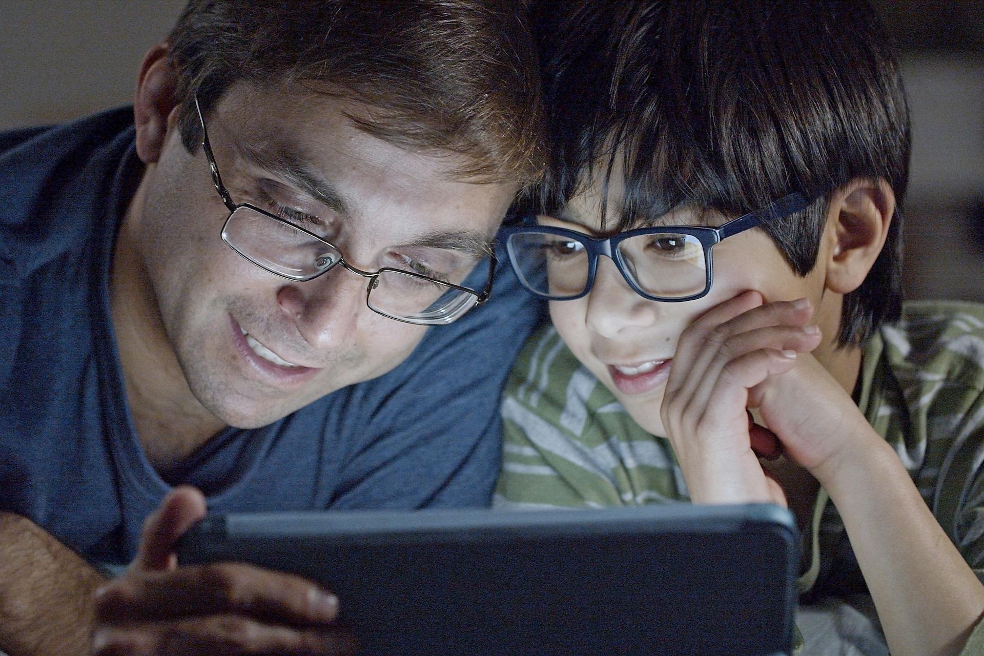 A father and son joyfully share a moment together, watching a tablet indoors.