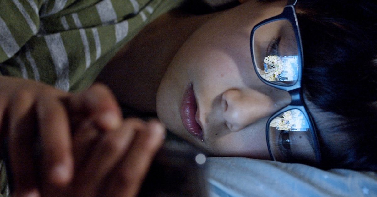A young person watching a movie on a laptop in a dark room, with a phone visibly face-down