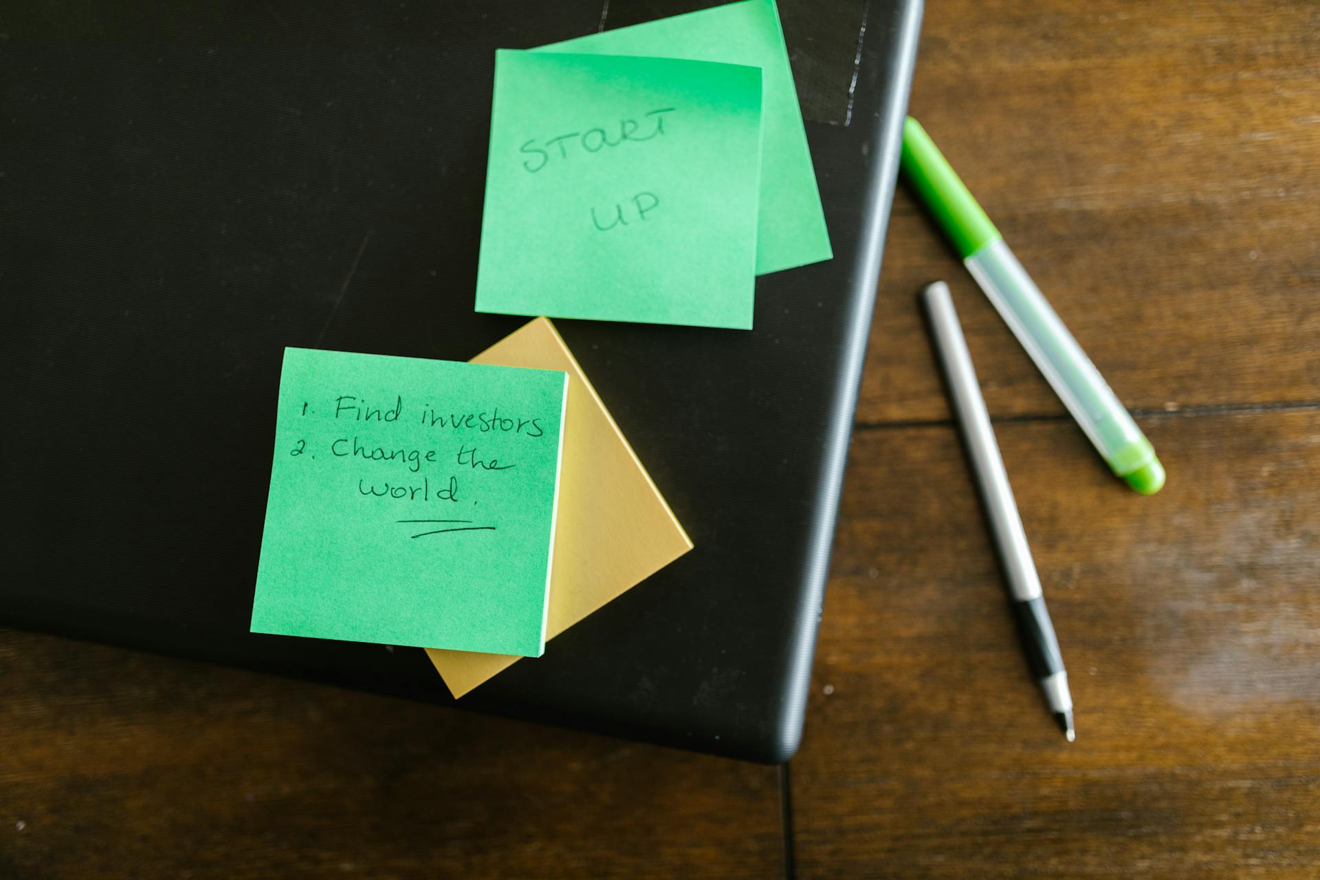 Green sticky notes with startup goals on a wooden desk with pens.