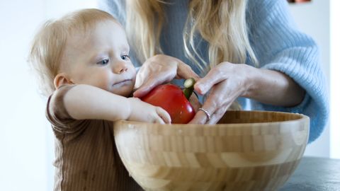 A parent helps a young child wash a red bell pepper in a wooden bowl, promoting healthy eating habits.