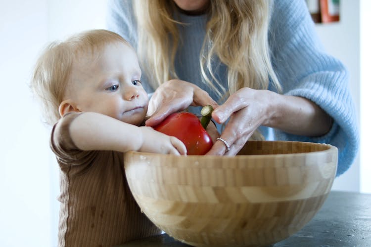 Woman Helping Toddler Putting Red Pepper On Wooden Bowl