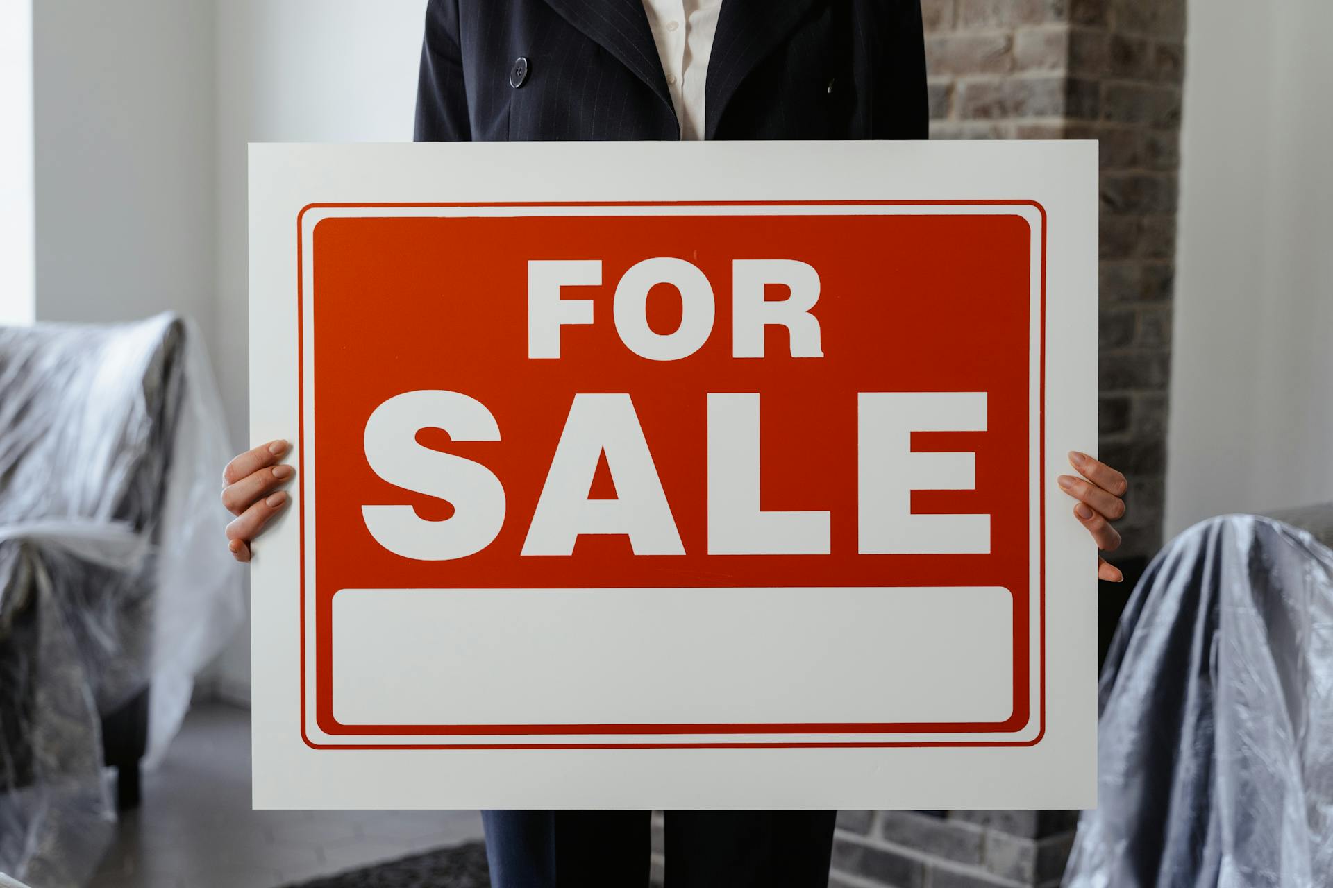 Close-up of a person in a suit holding a For Sale sign in a furnished room.