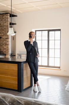 Professional woman in a black suit talking on a phone in a stylish kitchen interior.