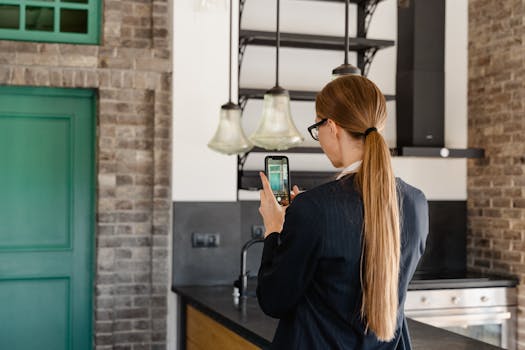 Rear view of a woman with long hair taking a photo with a smartphone in a stylish kitchen.