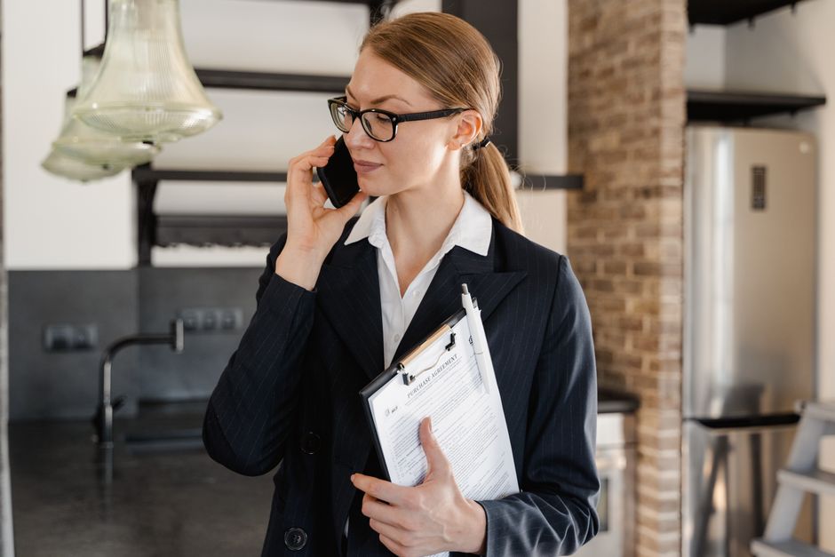 Empresária em um blazer preto utilizando um celular e segurando uma prancheta em um ambiente de escritório.