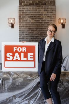 Businesswoman in suit with glasses holding a 'For Sale' sign indoors against a brick wall.