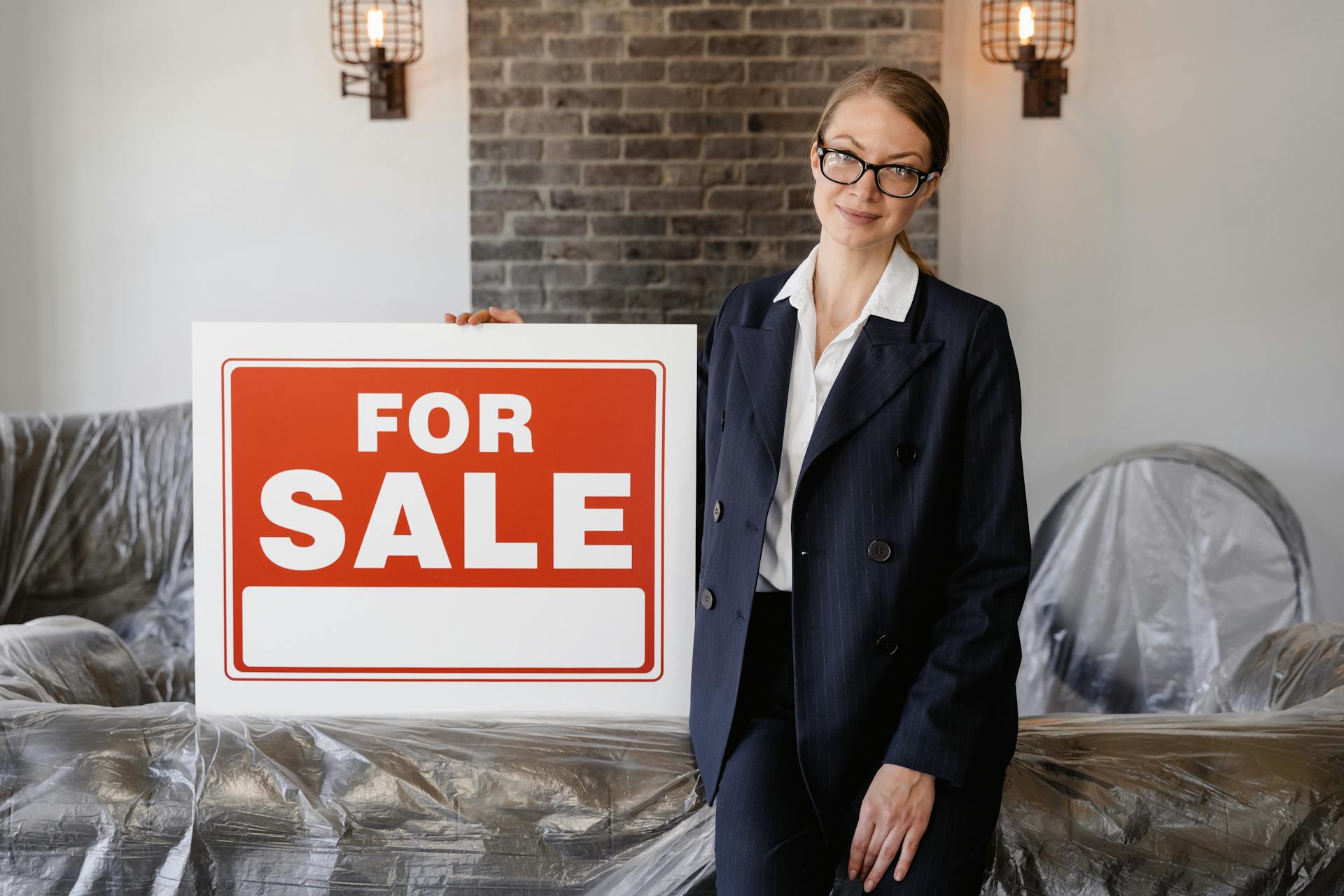 Real estate agent in formal attire holding a blank 'For Sale' sign inside an apartment.