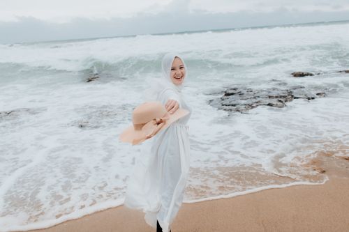 Free A smiling woman in a hijab enjoying the beach with waves and a straw hat in hand. Stock Photo