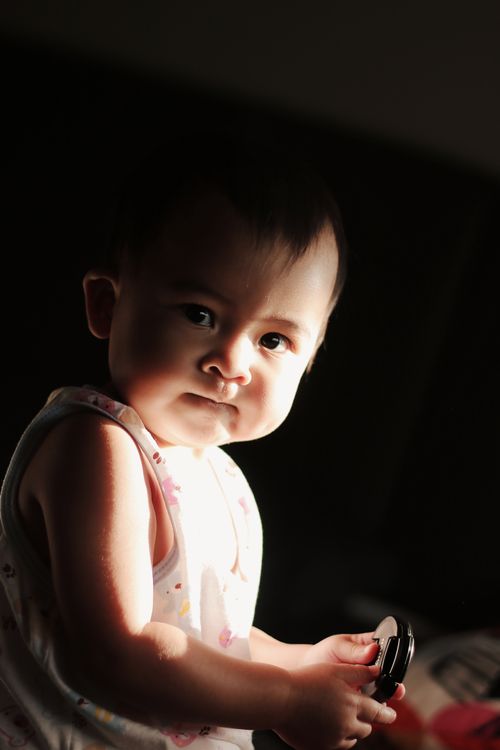 Free A cute baby with black hair holding an object, illuminated by warm sunlight indoors. Stock Photo