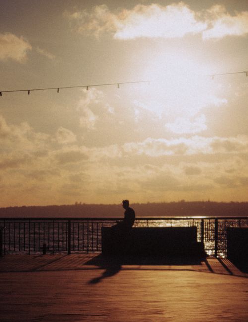 Free Silhouette of distant unrecognizable male siting on waterfront near metal railing and rippling river against cloudy sunset sky in town Stock Photo